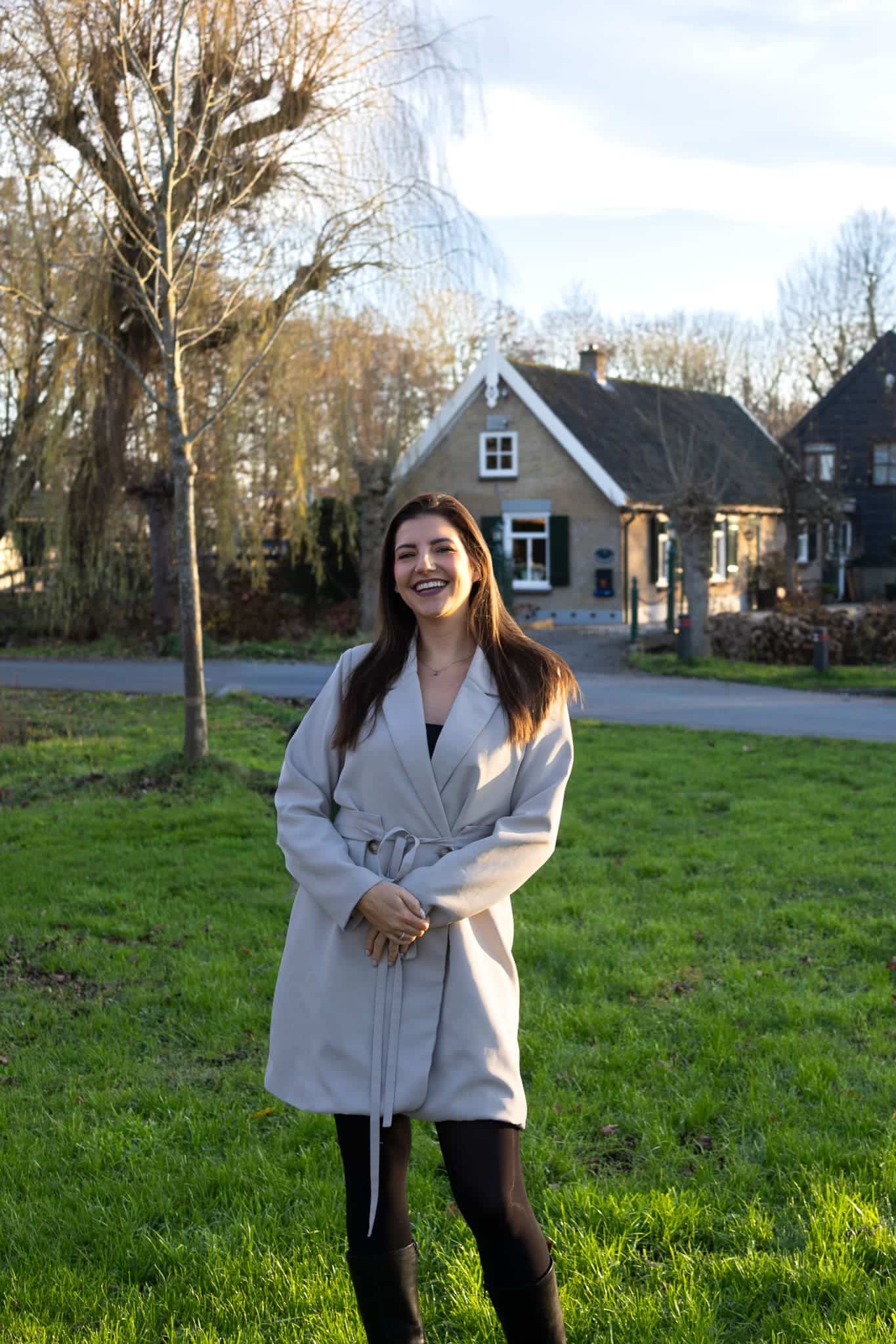 BURO210 Fullservice Marketingbureau Portret van een stralende vrouw, mogelijk een van onze specialisten, poserend in een landelijke omgeving met een charmant huis op de achtergrond. Ze lacht vriendelijk.