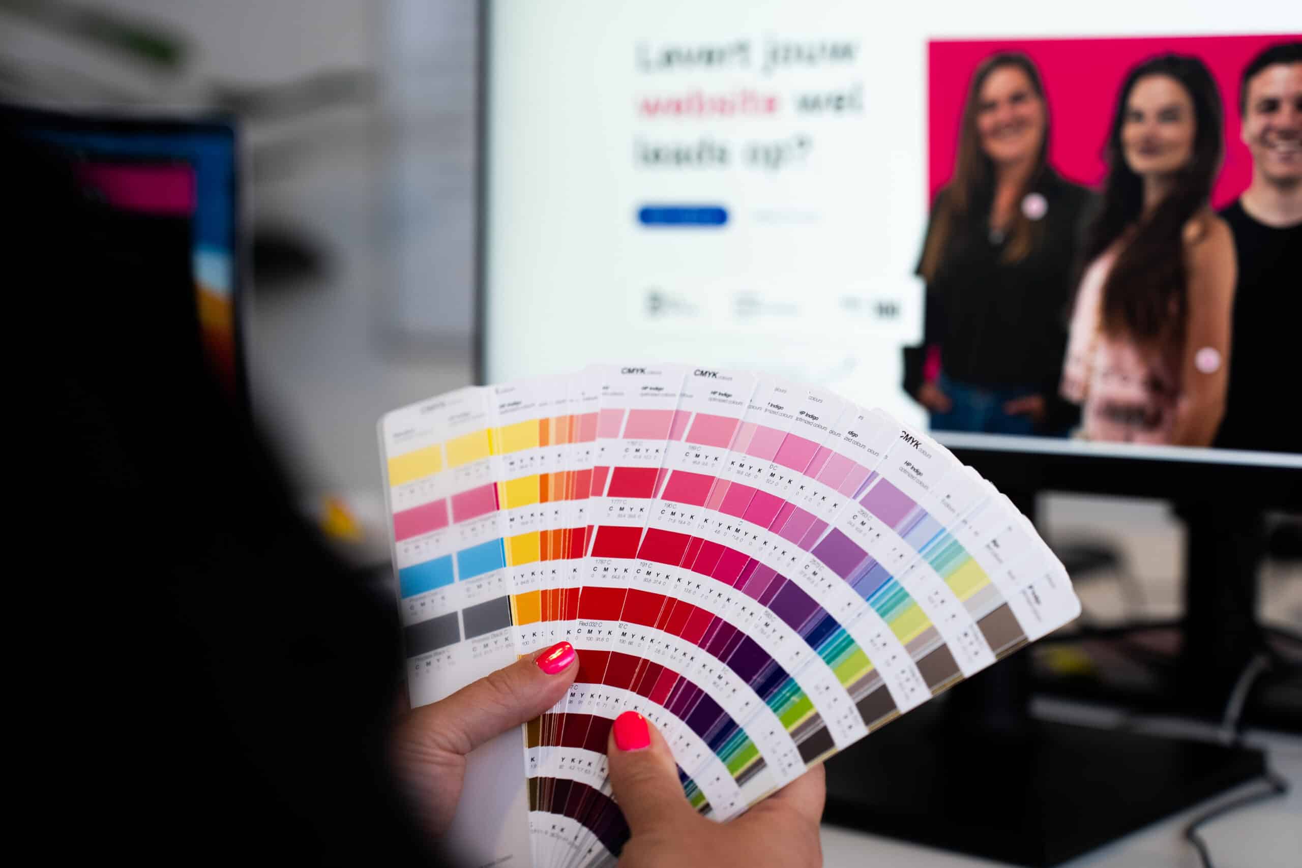 BURO210 Fullservice Marketingbureau Een vrouw houdt een CMYK kleurenpalet vast, belangrijk voor de visuele identiteit van een bedrijf. Op de achtergrond staat een computerscherm.