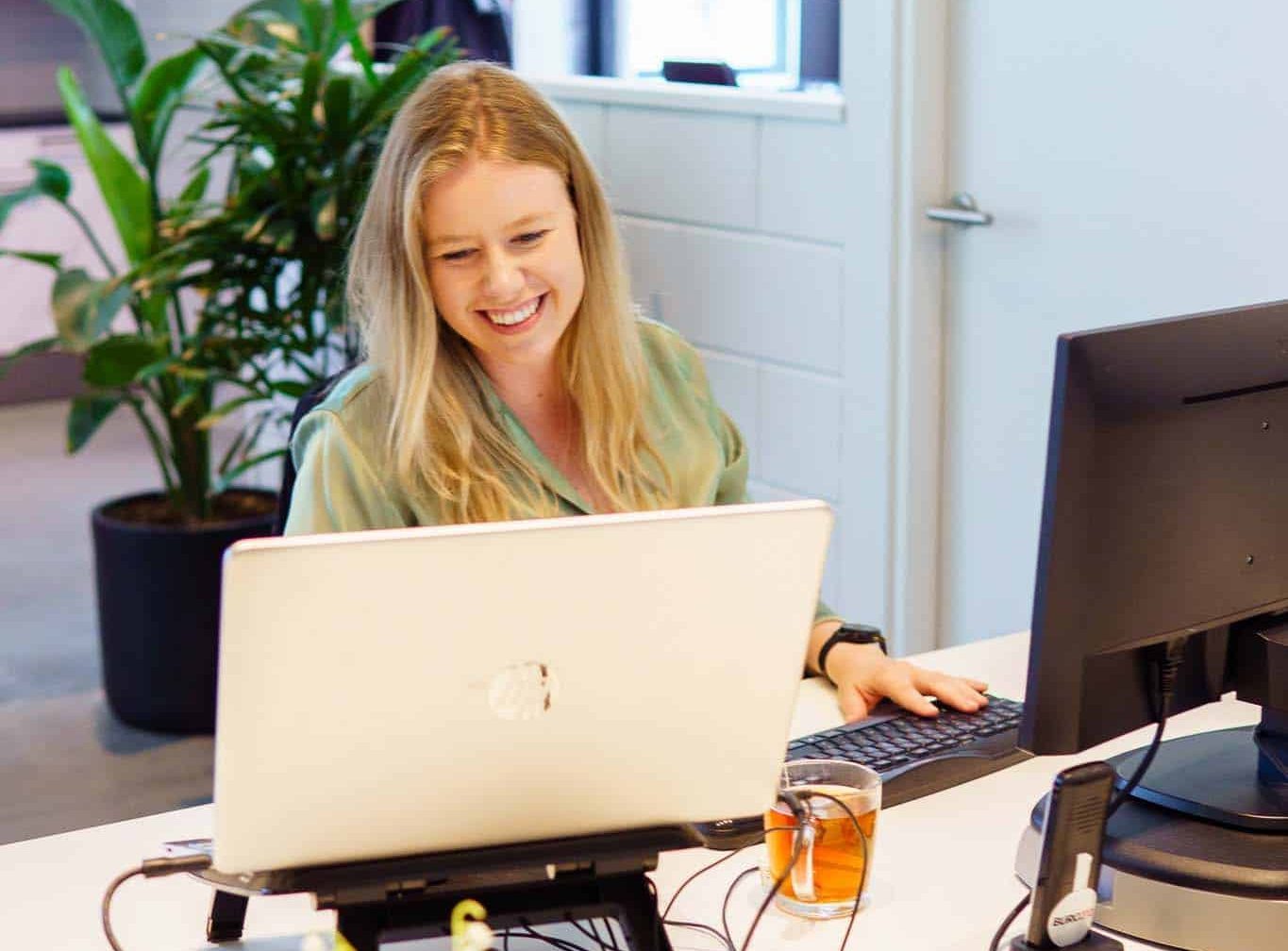 BURO210 Fullservice Marketingbureau Vrolijke administratief medewerker werkt aan de computer in een licht kantoor. Blonde vrouw typt op het toetsenbord, met een kop thee en planten op het bureau. Een prettige werkomgeving.