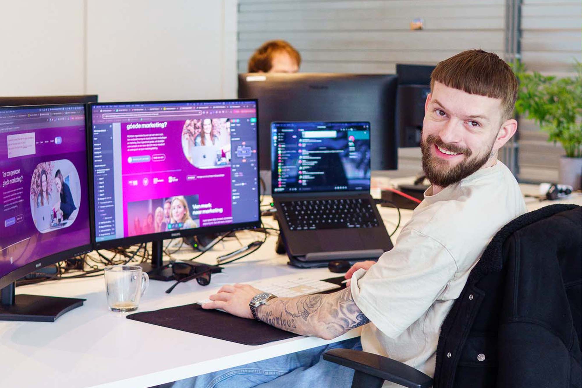 BURO210 Fullservice Marketingbureau Portret van een enthousiaste webdeveloper aan zijn bureau met computerschermen en een laptop. Hij glimlacht en kijkt naar de camera, bezig met webdevelopment.
