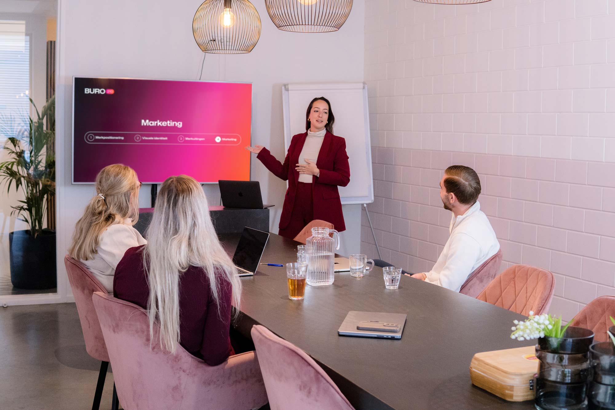 BURO210 Fullservice Marketingbureau Een jonge vrouw geeft een presentatie over marketing aan een groep mensen in een moderne kantooromgeving. Zoek naar een Traineeship Online Marketeer.