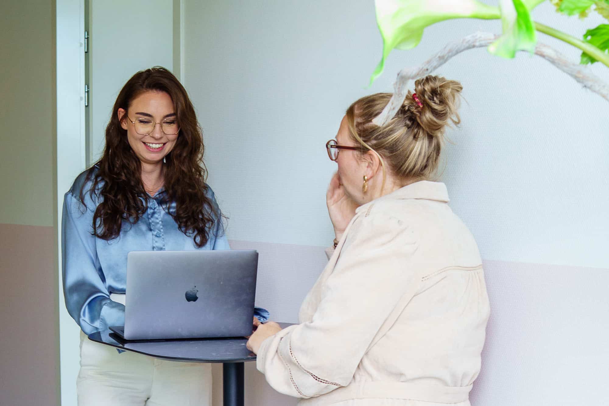 BURO210 Fullservice Marketingbureau Twee vrouwen werken samen aan een project. De een, mogelijk een Junior Projectmanager, gebruikt een laptop en lacht. Zakelijke sfeer, lichte kleuren.