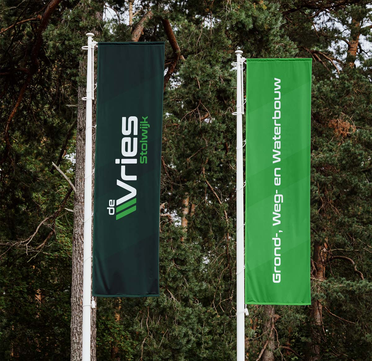 BURO210 Fullservice Marketingbureau Twee vlaggen van De Vries Stolwijk, een donkergroene en een lichtgroene, wapperend voor bomen. Perfect voor grond-, weg- en waterbouw projecten.